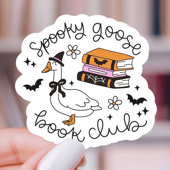 A hand holds a die-cut sticker featuring a white goose wearing a black witch hat and bow, with the words 'Spooky Goose Book Club' in black script. The design also includes three stacked books in orange, pink, and black with bat and flower illustrations, against a blurred indoor background.