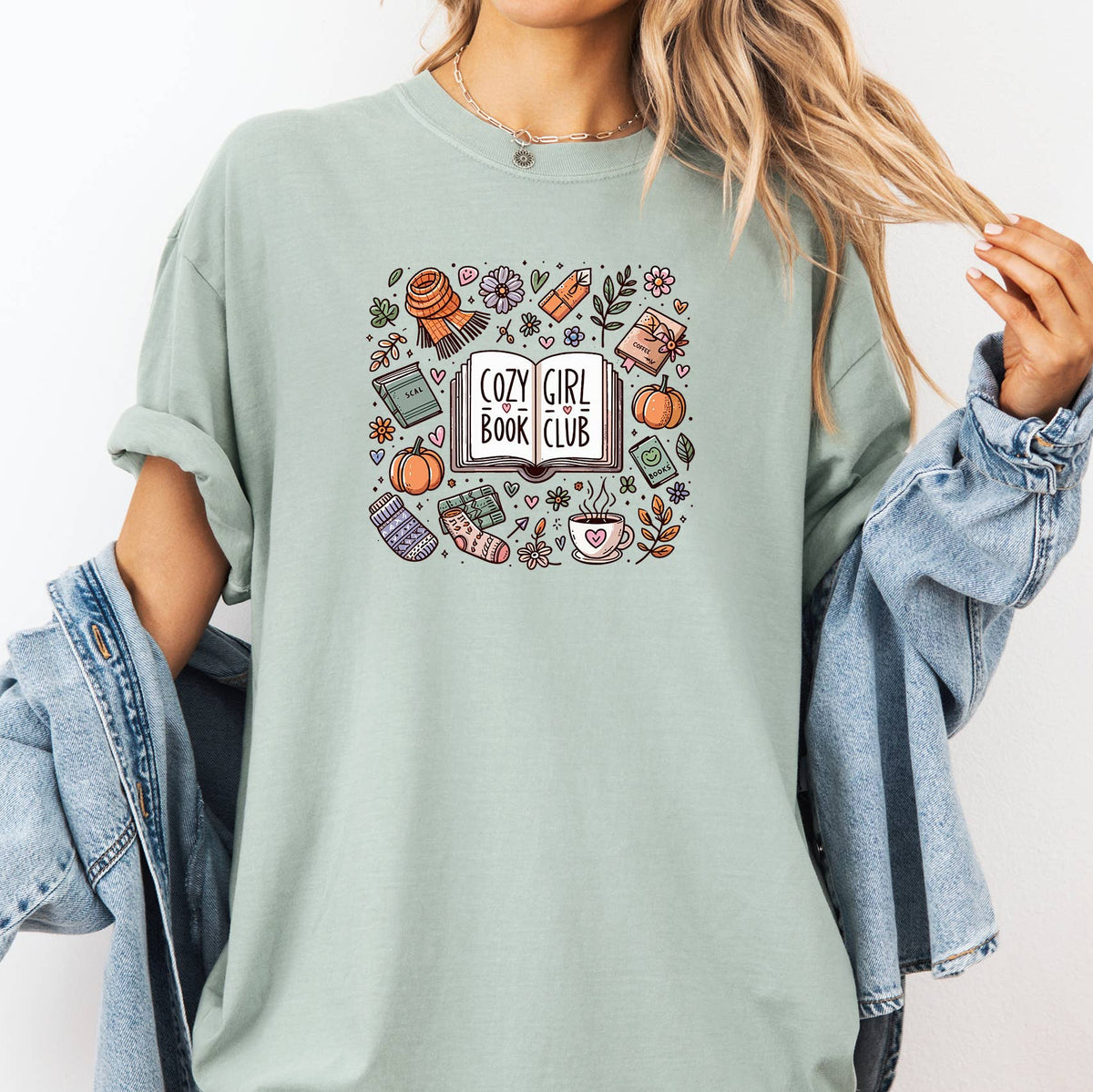 A woman wearing a sage green graphic t-shirt with a 'Cozy Girl Book Club' illustration, styled with a light blue denim jacket draped off her shoulders against a plain white background.