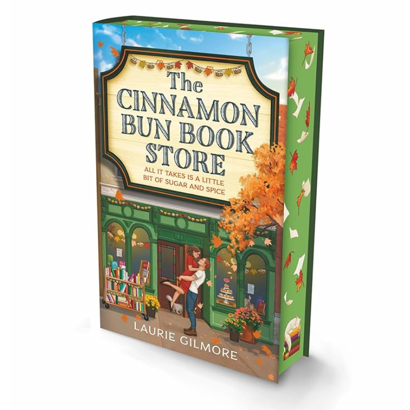 The Cinnamon Bun Book Store paperback book by Laurie Gilmore, shown at a three-quarter angle against a plain white background. The cover features a colorful illustration of a cozy bookshop with autumn foliage and a couple embracing outside.
