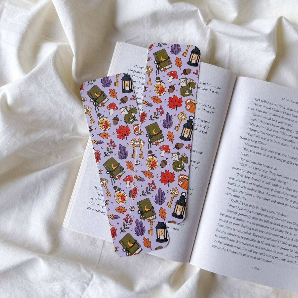 Two bookmarks with a whimsical autumn pattern of pumpkins, lanterns, acorns, and leaves on a lavender background, resting on an open book over a draped white fabric background.