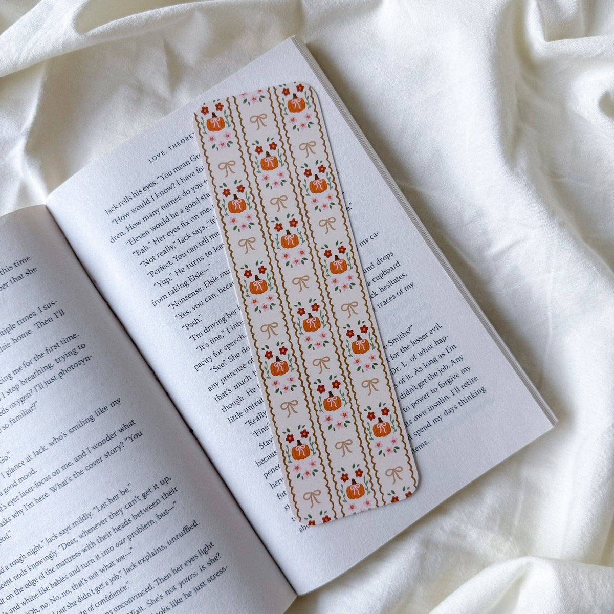 A decorative paper bookmark with a repeating pumpkin and bow pattern on a cream background, placed on an open book resting on draped white fabric.