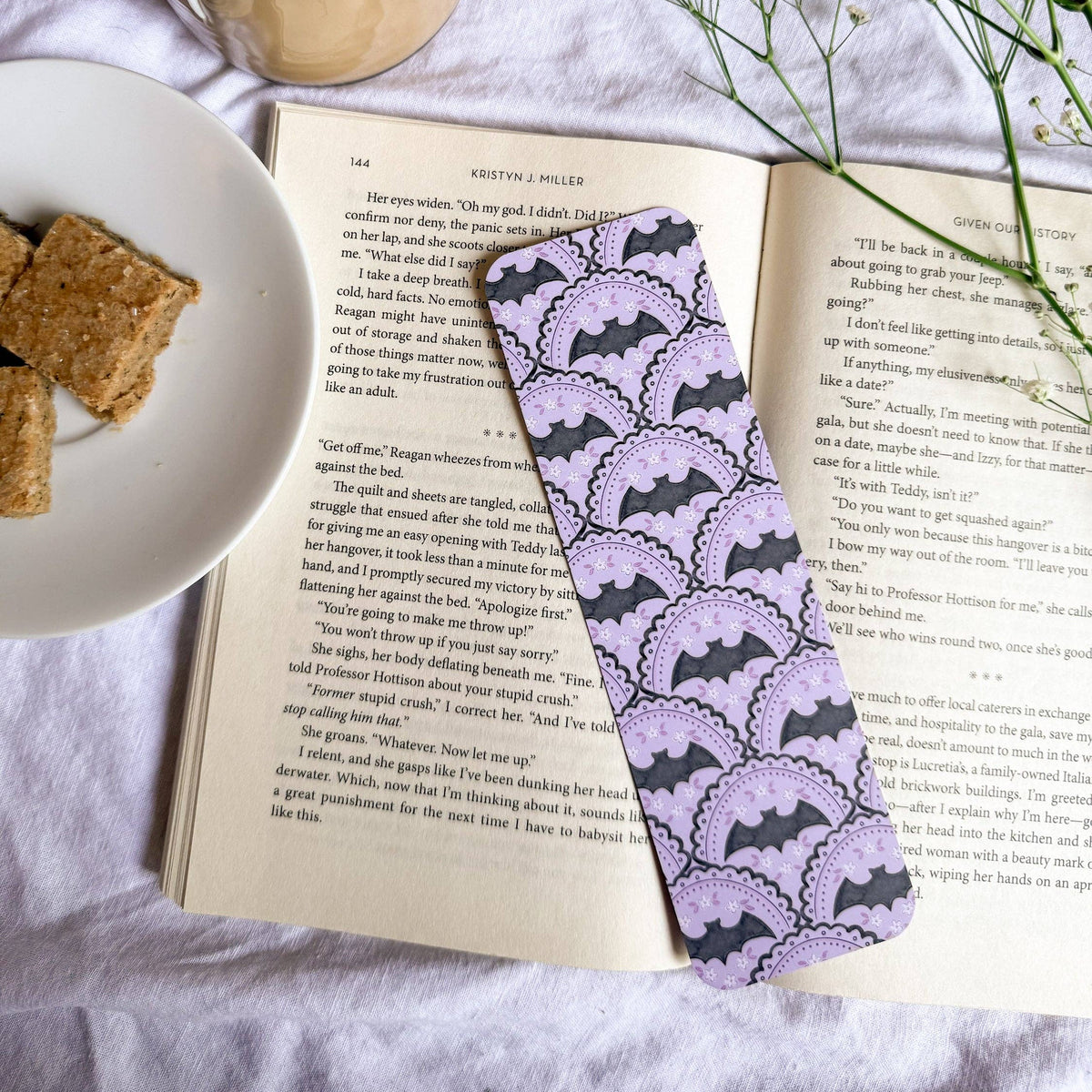 Open book with a purple lace bookmark, cup of coffee, and cookies on a light surface.