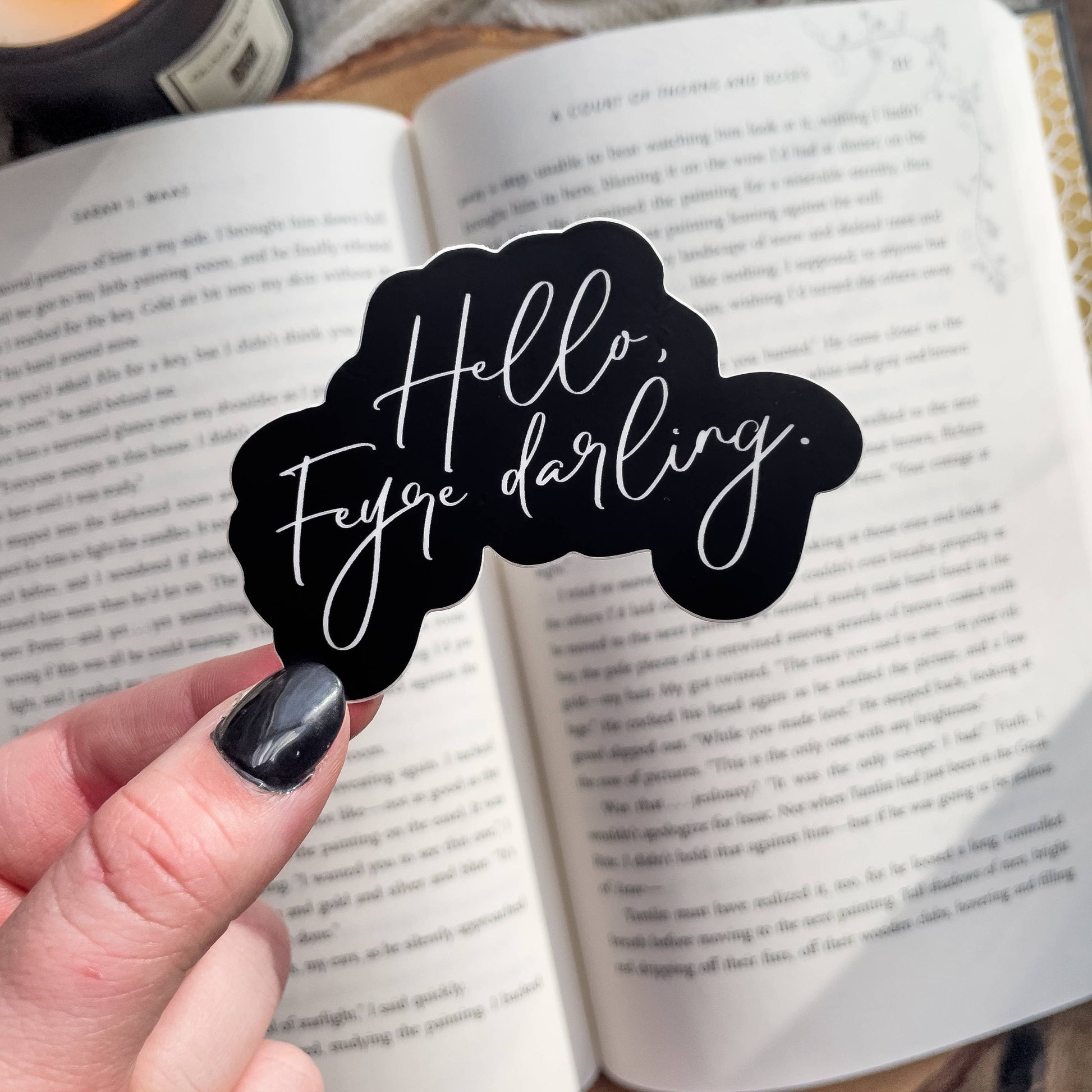 Black sticker with 'Hello, Feyre darling' text held over an open book.