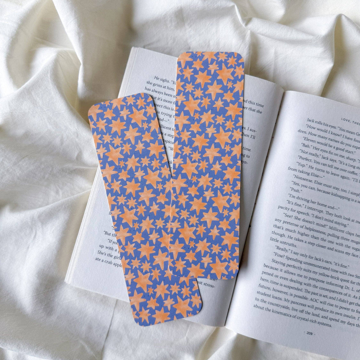 Two paper bookmarks with a blue and orange floral pattern, resting on an open book over a draped white fabric background.