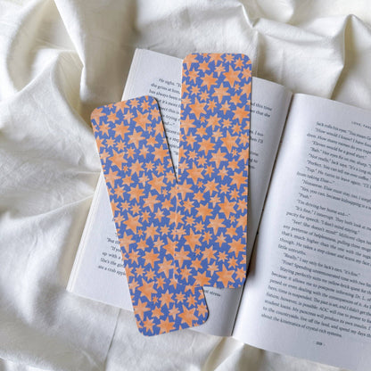 Two paper bookmarks with a blue and orange floral pattern, resting on an open book over a draped white fabric background.
