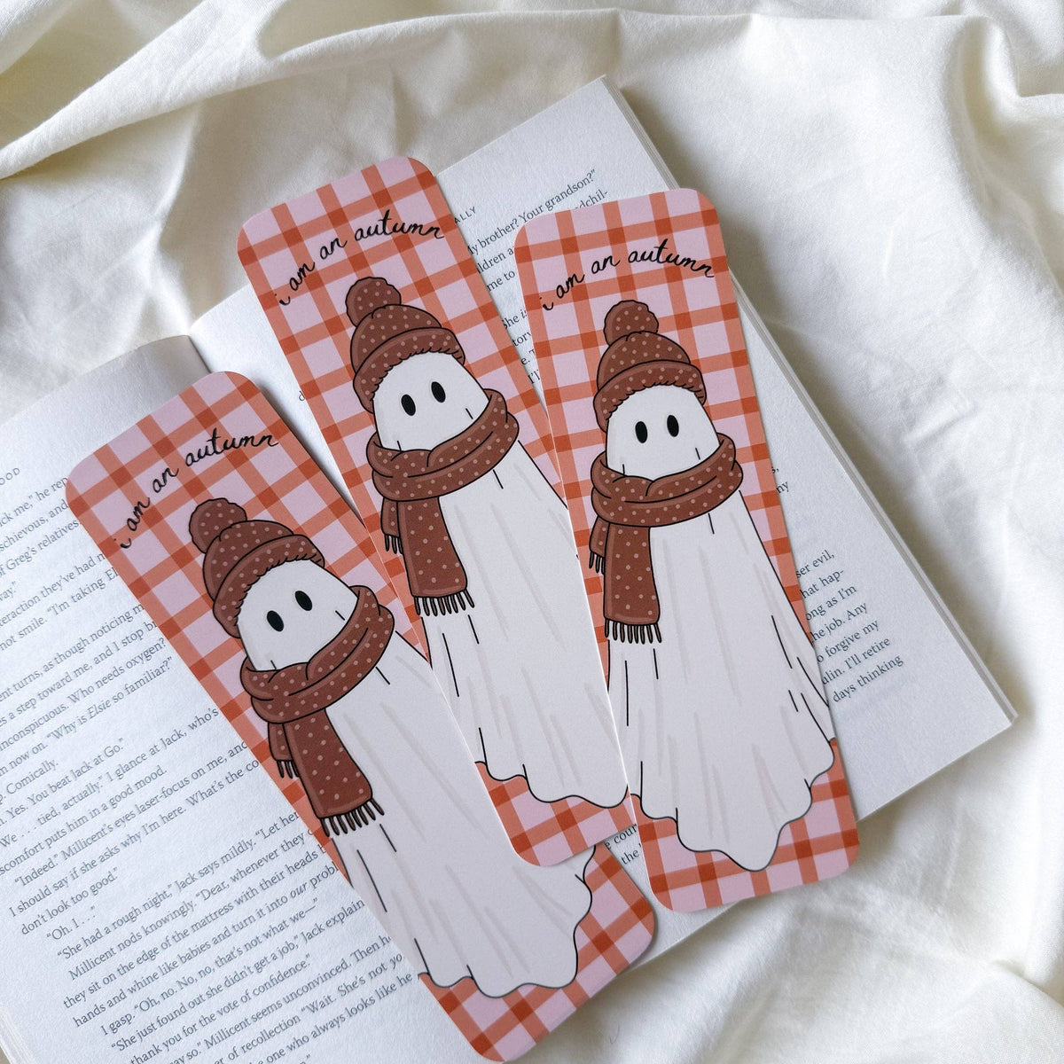 Three ghost-shaped bookmarks with a red and white gingham pattern, brown knit hats, and brown knit scarves, resting on an open book over a draped white fabric background.