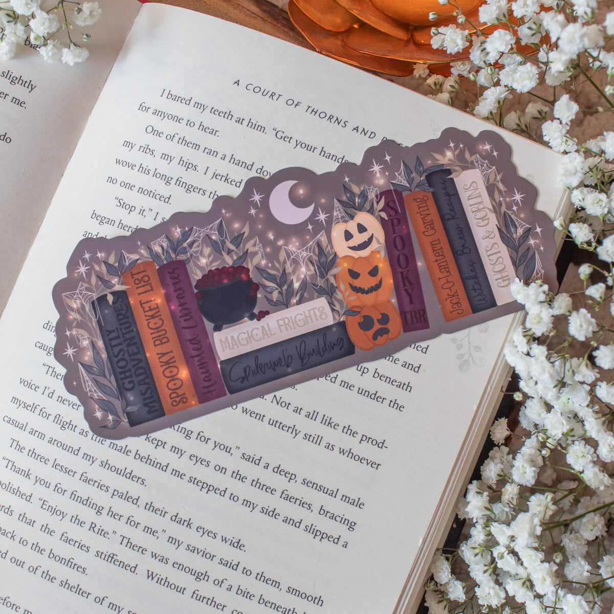 A decorative book bookmark styled like a row of vintage books with Halloween-themed accents, resting on an open book surrounded by white baby's breath flowers and a glowing orange pumpkin ornament.