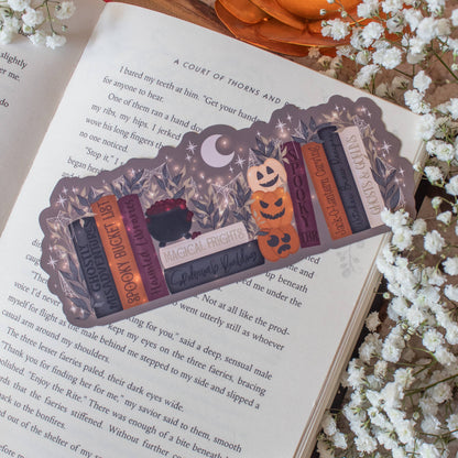 A decorative book bookmark styled like a row of vintage books with Halloween-themed accents, resting on an open book surrounded by white baby's breath flowers and a glowing orange pumpkin ornament.