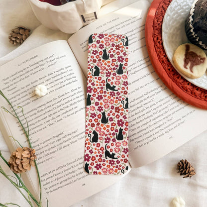 A rectangular book bookmark with a red, pink, and orange floral pattern and black cat silhouettes, resting on an open book. The scene is styled with a white mug of tea, pinecones, and a plate of cookies on a white fabric background.