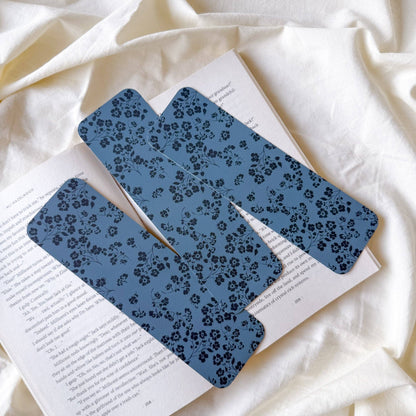 Two blue bookmarks with a black floral pattern resting on an open book, set against a background of draped cream-colored fabric.