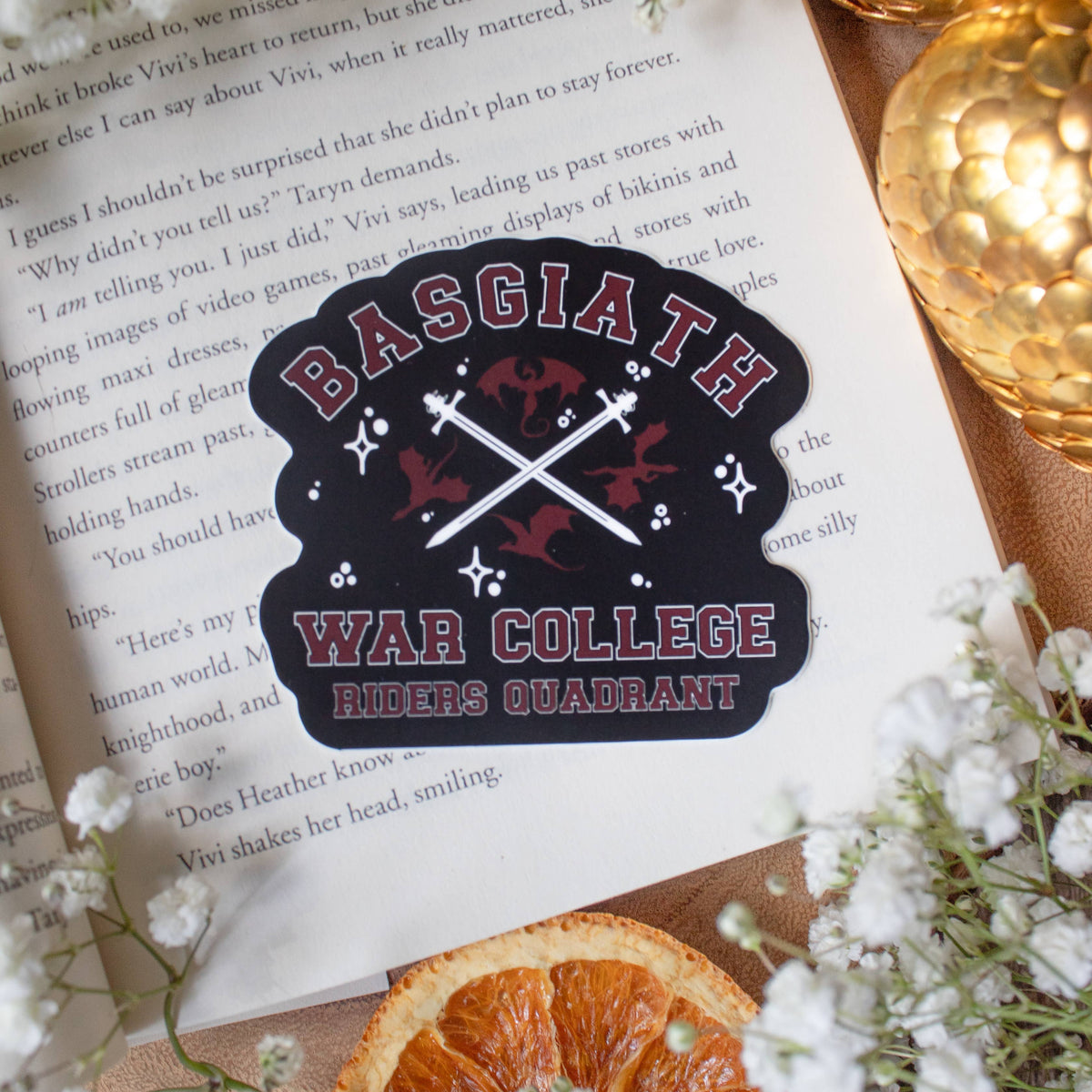 A die-cut sticker featuring the Basgiath War College Riders Quadrant from The Godfather, styled on top of an open book with dried orange slices and white flowers.