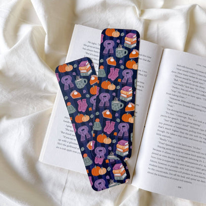 Two patterned book bookmarks with purple, orange, and teal illustrations of pumpkins, books, and coffee cups, resting on an open book over a draped white fabric background.
