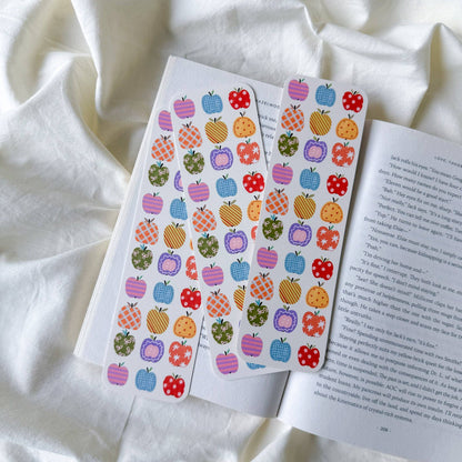 Two sheets of colorful apple-shaped stickers with various patterns (solid colors, stripes, polka dots) laid out on an open book over a draped white fabric background.