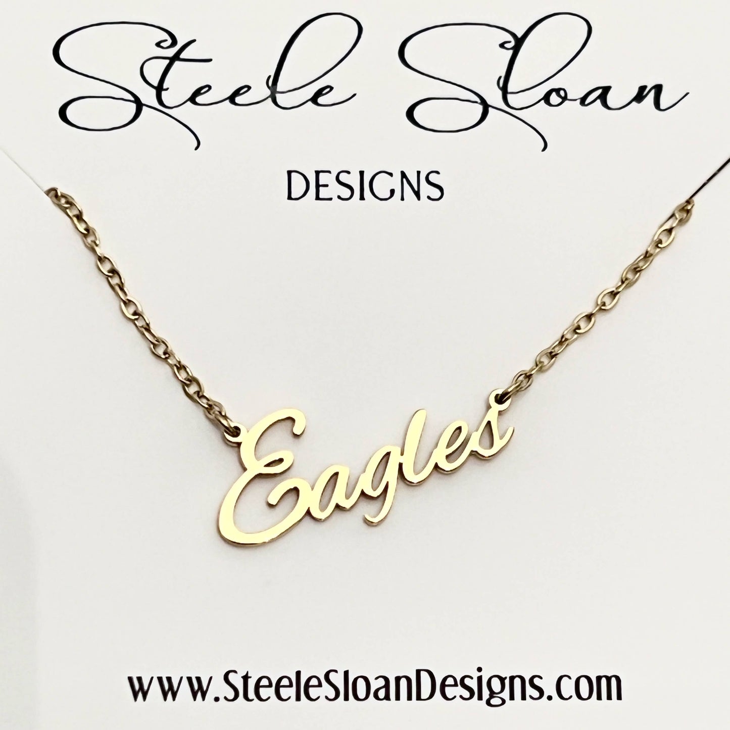 A gold nameplate necklace spelling 'Eagles' on a delicate gold chain, presented on a white display card with the brand name 'Steele Sloan DESIGNS' at the top and the website 'www.SteeleSloanDesigns.