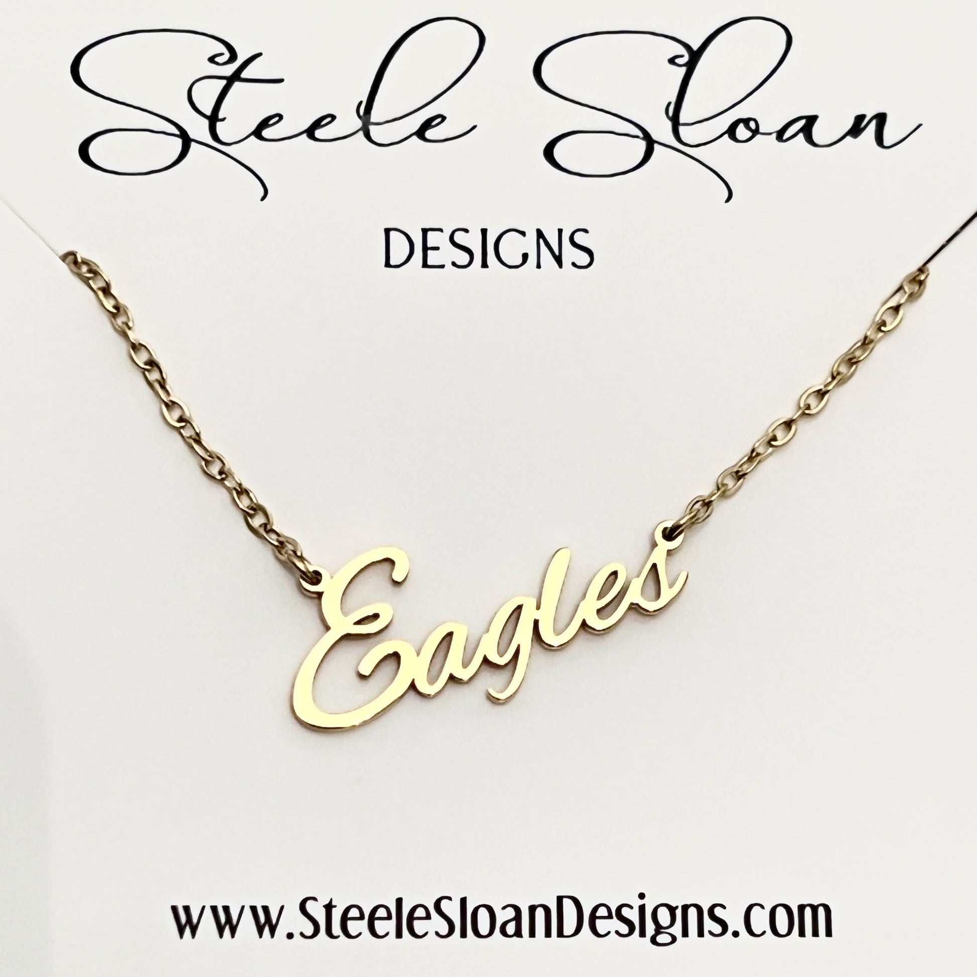 A gold nameplate necklace spelling 'Eagles' on a delicate gold chain, presented on a white display card with the brand name 'Steele Sloan DESIGNS' at the top and the website 'www.SteeleSloanDesigns.
