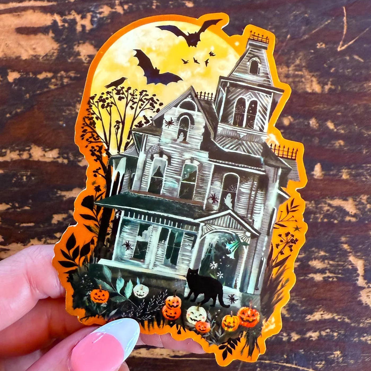 Halloween-themed sticker of a haunted house with pumpkins and bats on a wooden surface.