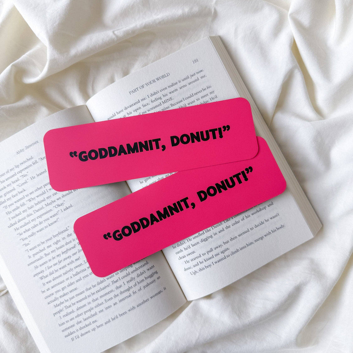 Two bright pink rectangular bookmarks with black text reading “GODDAMNIT, DONUT!” placed on an open book laid on white fabric.