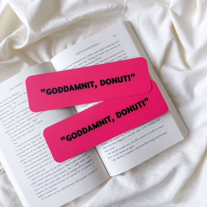 Two bright pink rectangular bookmarks with black text reading “GODDAMNIT, DONUT!” placed on an open book laid on white fabric.