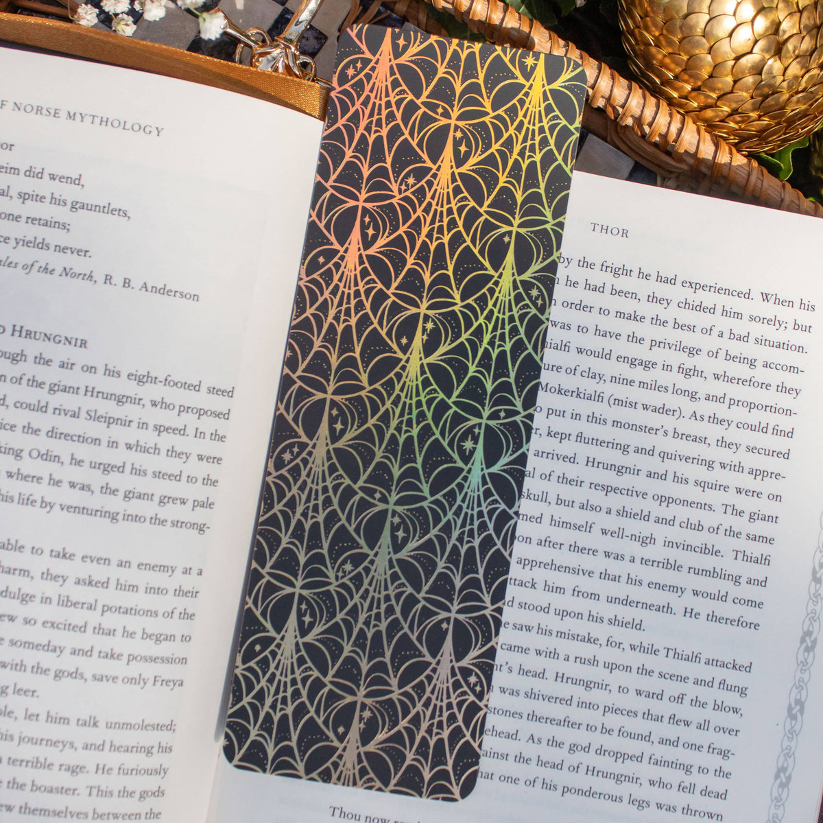 A rectangular black bookplate with an intricate gold spiderweb pattern, pinned to the pages of an open book. The scene is styled with a woven basket, green ivy, and a gold pinecone on a white background.