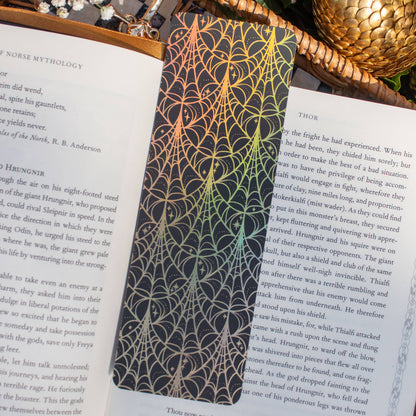 A rectangular black bookplate with an intricate gold spiderweb pattern, pinned to the pages of an open book. The scene is styled with a woven basket, green ivy, and a gold pinecone on a white background.