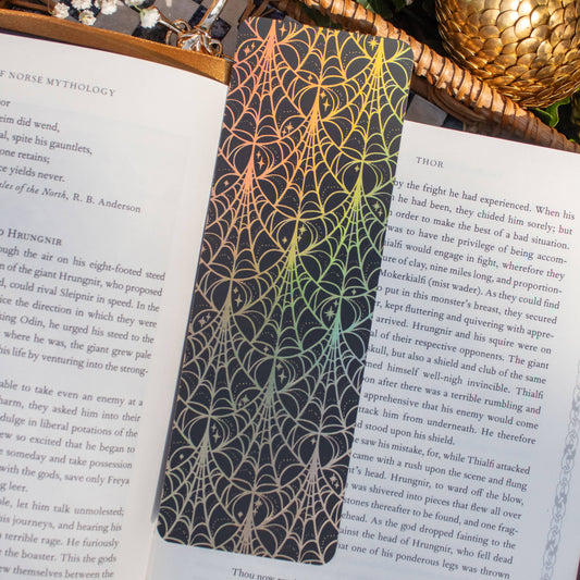 A rectangular black bookplate with an intricate gold spiderweb pattern, pinned to the pages of an open book. The scene is styled with a woven basket, green ivy, and a gold pinecone on a white background.