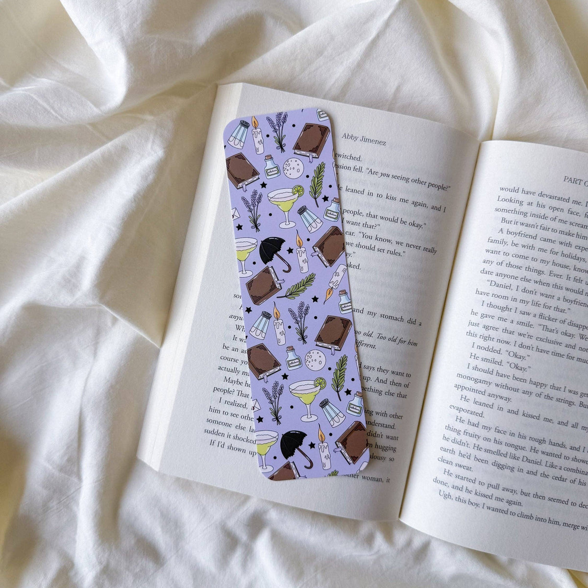 A lavender bookmark with a coffee-themed pattern, resting on an open book over a soft white fabric background.
