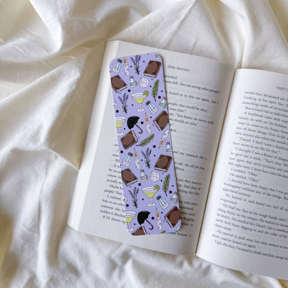 A lavender bookmark with a coffee-themed pattern, resting on an open book over a soft white fabric background.