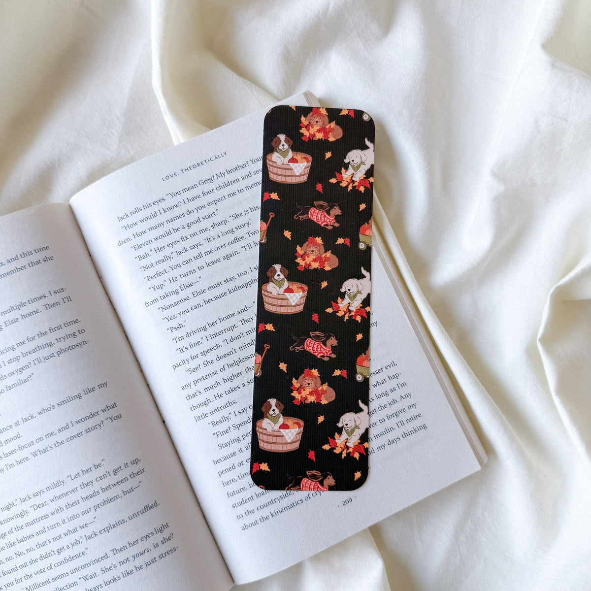 A slim black book bookmark with a whimsical pattern of white rabbits, red roses, and layered cake slices, resting on an open book over white fabric.