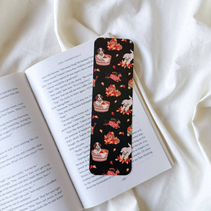 A slim black book bookmark with a whimsical pattern of white rabbits, red roses, and layered cake slices, resting on an open book over white fabric.