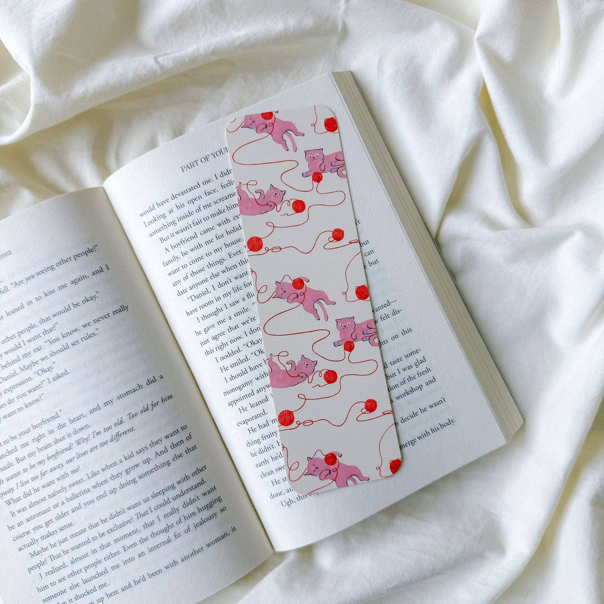 A decorative bookmark with a pink and red cat-and-ball pattern, shown resting on an open book page, all styled on a soft white fabric background.