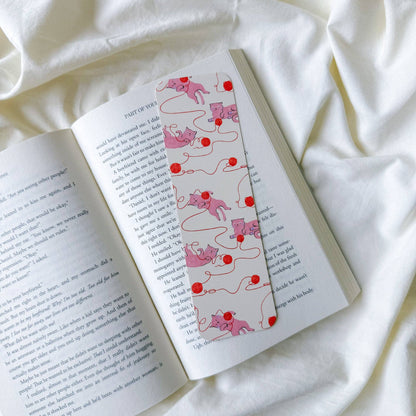 A decorative bookmark with a pink and red cat-and-ball pattern, shown resting on an open book page, all styled on a soft white fabric background.