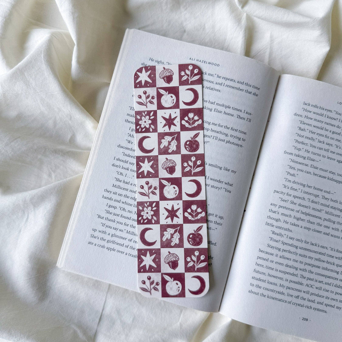 A decorative bookmark with a checkerboard pattern of autumn and winter motifs in dark red and cream, shown inside an open book on a draped white fabric background.