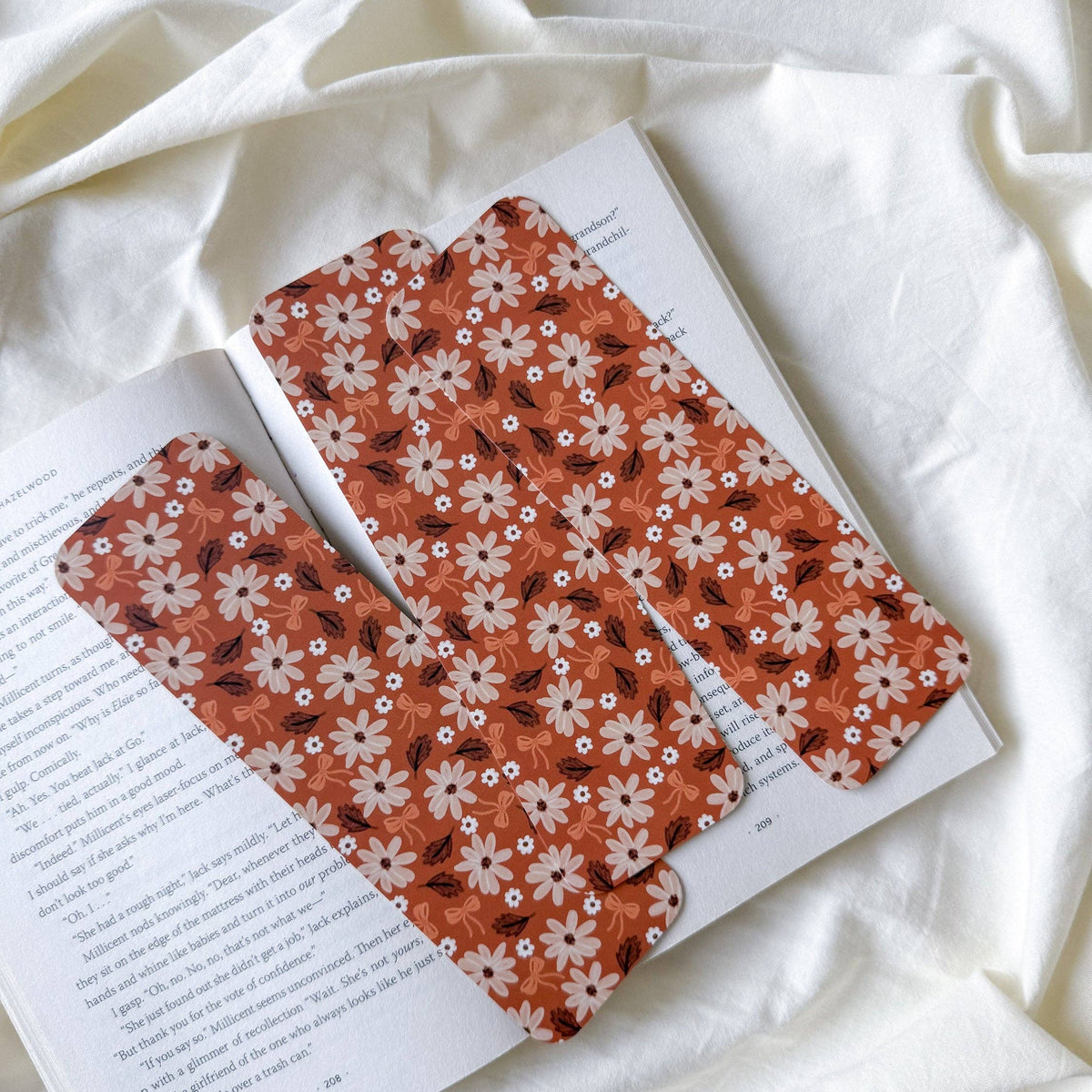 Two rectangular fabric bookmarks with a rust-orange background and a white floral pattern are laid on an open book over a draped white fabric background.