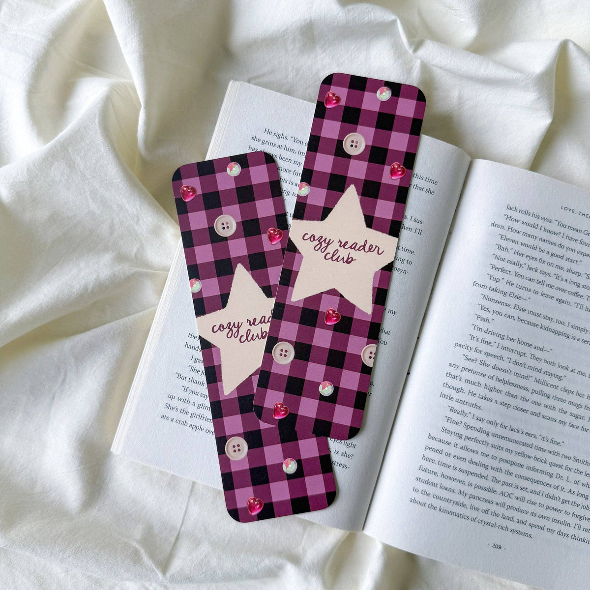 Two pink and black buffalo plaid bookmarks with white buttons and star-shaped tabs reading 'easy reader club' are placed on an open book over white fabric.