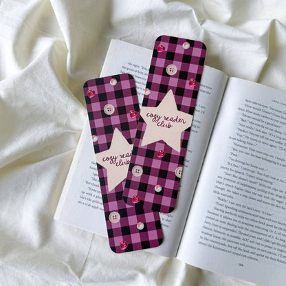 Two pink and black buffalo plaid bookmarks with white buttons and star-shaped tabs reading 'easy reader club' are placed on an open book over white fabric.