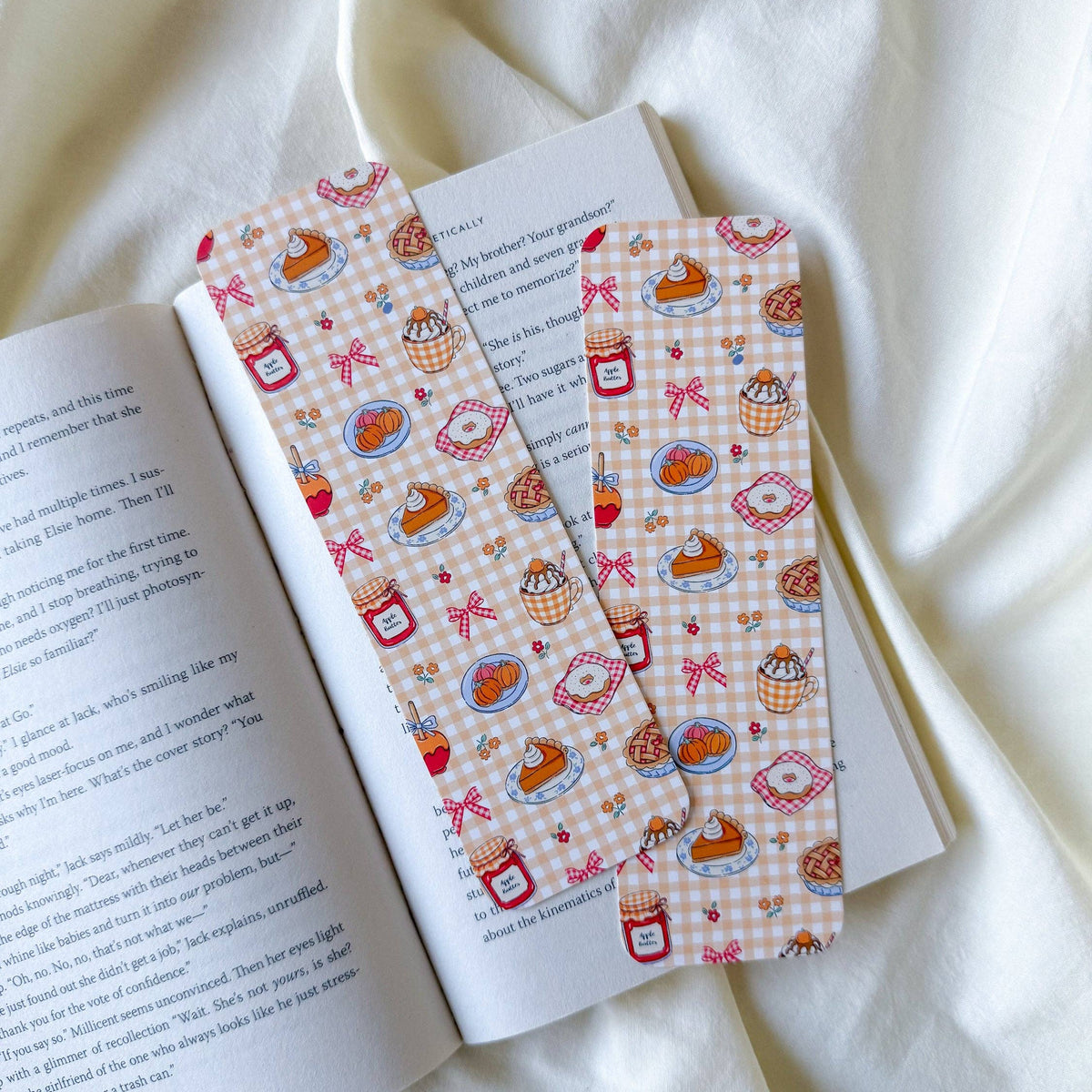 Two rectangular bookmarks with a pink and white gingham pattern featuring illustrations of cupcakes, coffee cups, and red bows, laid on an open book over a draped white fabric background.