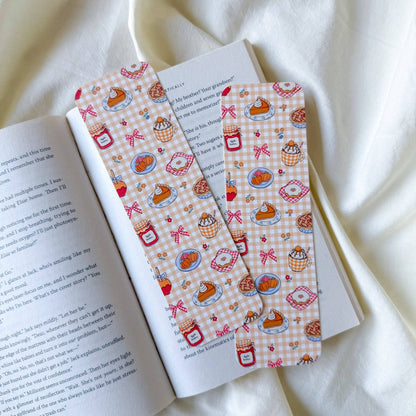 Two rectangular bookmarks with a pink and white gingham pattern featuring illustrations of cupcakes, coffee cups, and red bows, laid on an open book over a draped white fabric background.