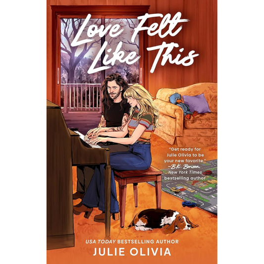Book cover for 'Love Fell Like This' by Julie Olivia, featuring an illustrated couple sitting together in a warmly lit living room with a piano, orange walls, and a dog on the floor.