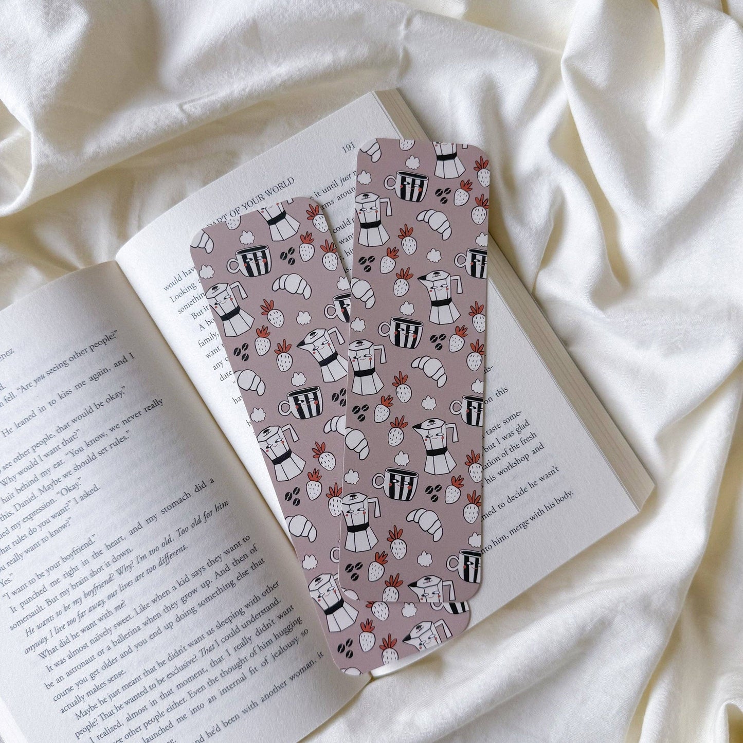 Two patterned bookmarks with a dusty pink background and black-and-white espresso cup illustrations, resting on an open book over a white fabric background.