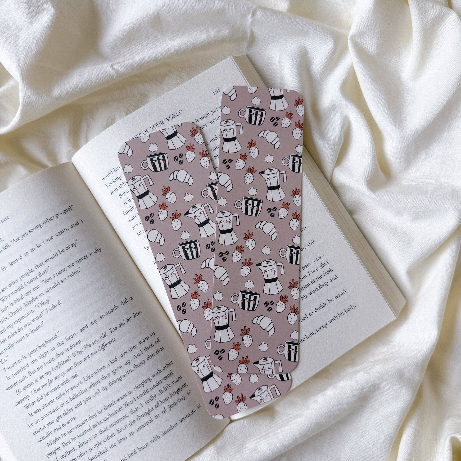 Two patterned bookmarks with a dusty pink background and black-and-white espresso cup illustrations, resting on an open book over a white fabric background.