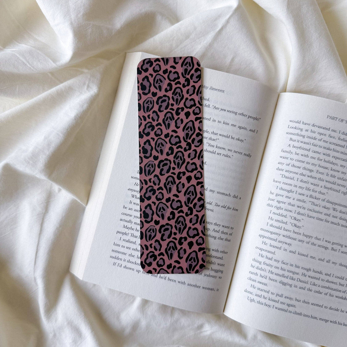A slim rectangular bookmark with a pink and black leopard print pattern, resting on an open book laid on white fabric.