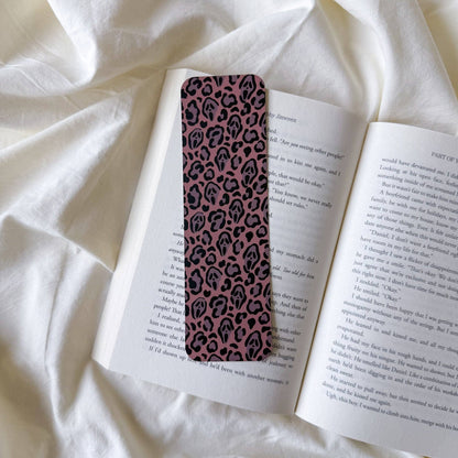 A slim rectangular bookmark with a pink and black leopard print pattern, resting on an open book laid on white fabric.