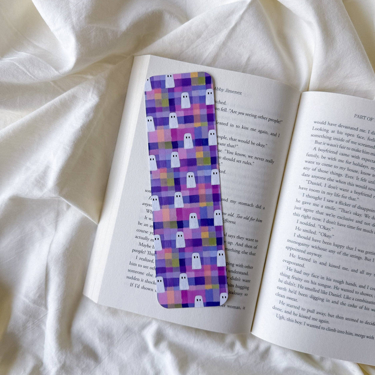 A rectangular book bookmark with a purple and pink pixelated ghost pattern, placed on an open book on a white fabric background.