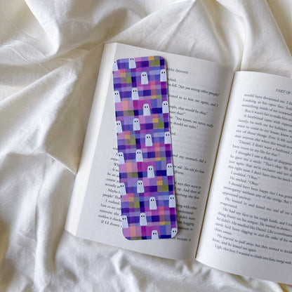 A rectangular book bookmark with a purple and pink pixelated ghost pattern, placed on an open book on a white fabric background.