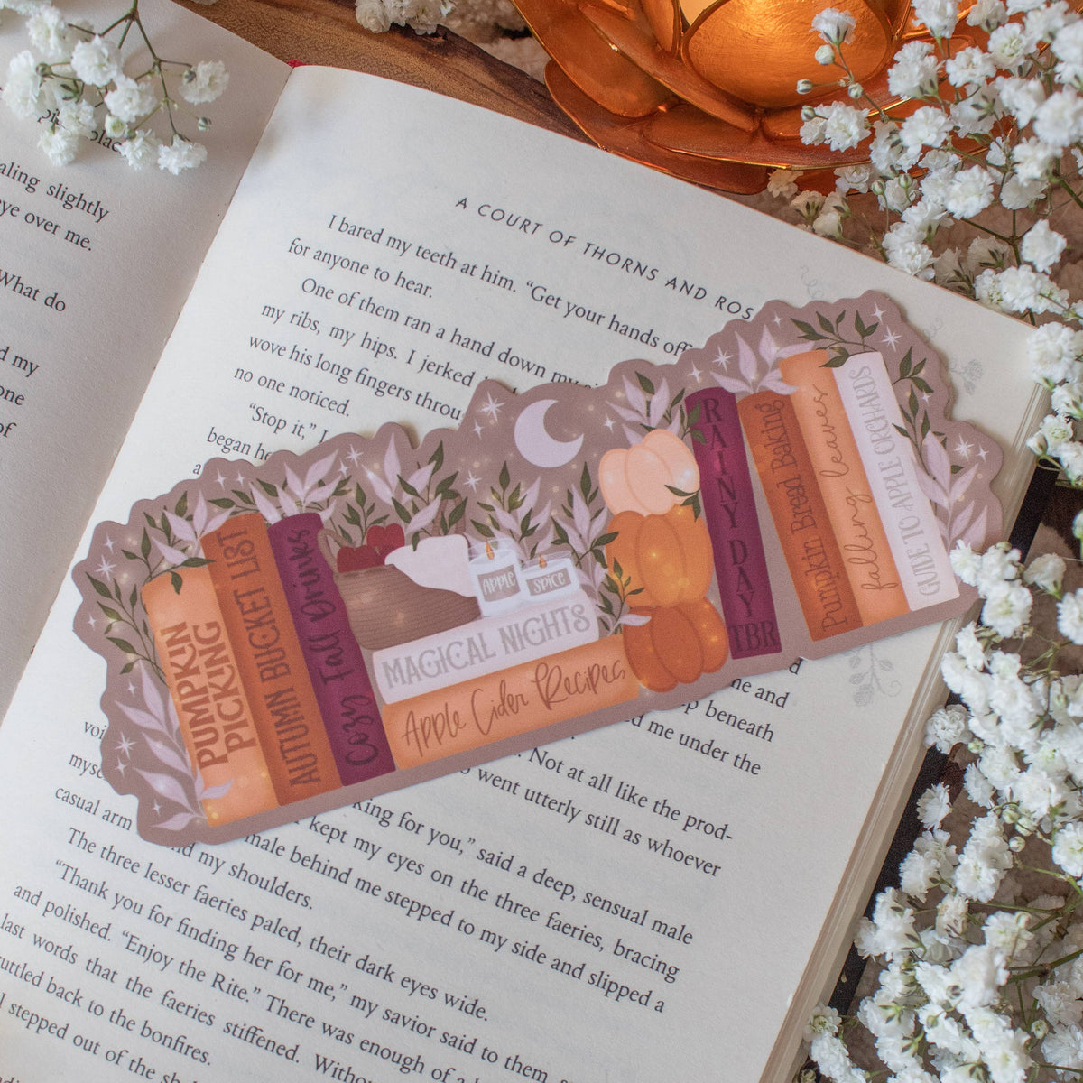 A decorative book bookmark styled on an open book surrounded by white baby's breath flowers and an orange pumpkin. The bookmark features a vintage illustration of a woman in a pink dress and a stack of colorful books with text like 'MAGICAL NIGHTS' and 'OLD LOVE REWIND'.